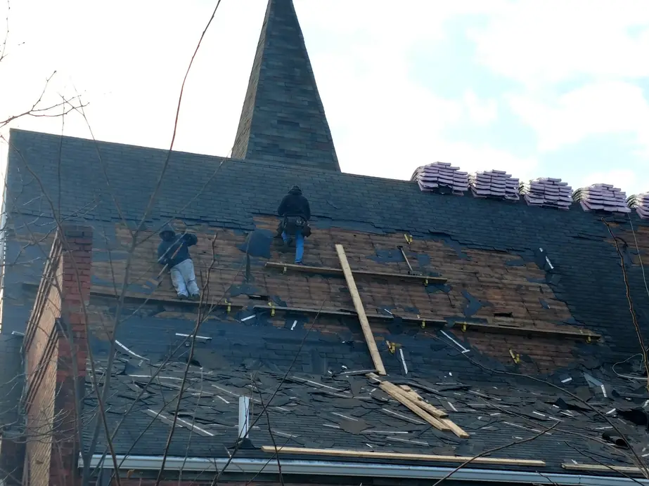 Two roofers on the steep church roof mid tear-off, new shingle bundles along the ridge